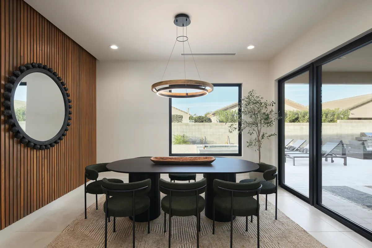 A bright dining area featuring a round table with chic chairs, a large mirror reflecting natural light, and a warm, welcoming ambiance. The setting is ideal for group dinners and celebrations.