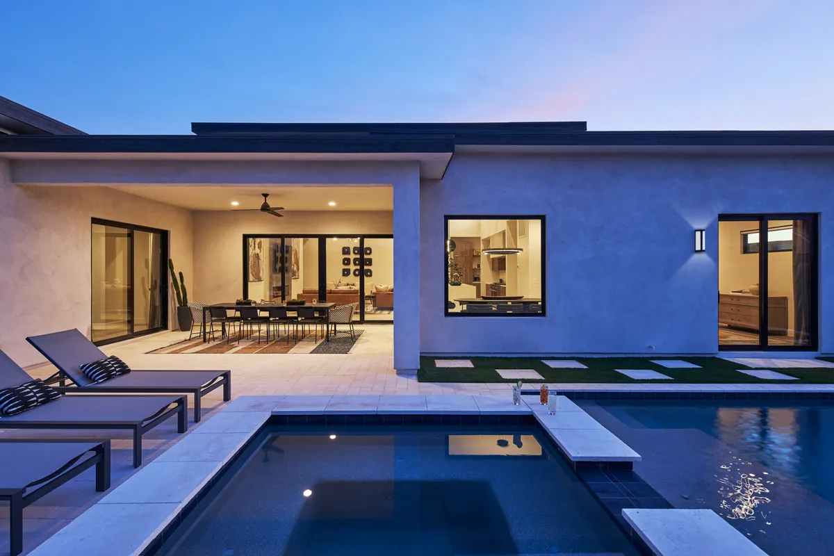 The image showcases a stunning modern backyard at sunset, featuring a sparkling private pool surrounded by sleek lounge chairs, an inviting outdoor dining area, and a glimpse into a chic, open-concept interior living space. The scene is set for relaxation, festive group gatherings, and memorable evenings under the Arizona sky.