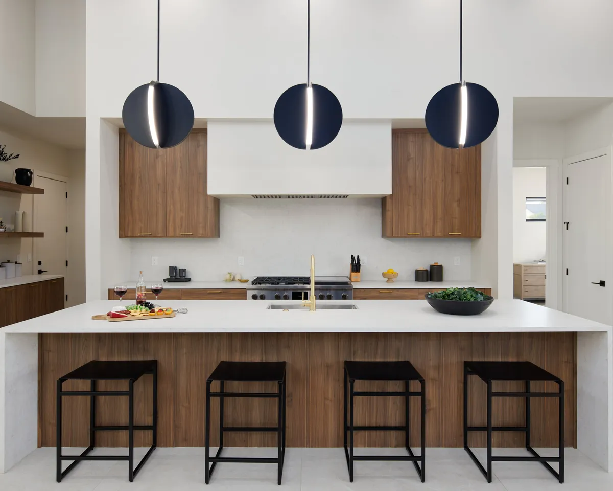 A spacious, modern kitchen with a colossal island countertop, pendant lighting, sleek wood cabinetry, and stools. The kitchen is set up for both cooking and entertaining, with a food platter suggesting culinary activity.