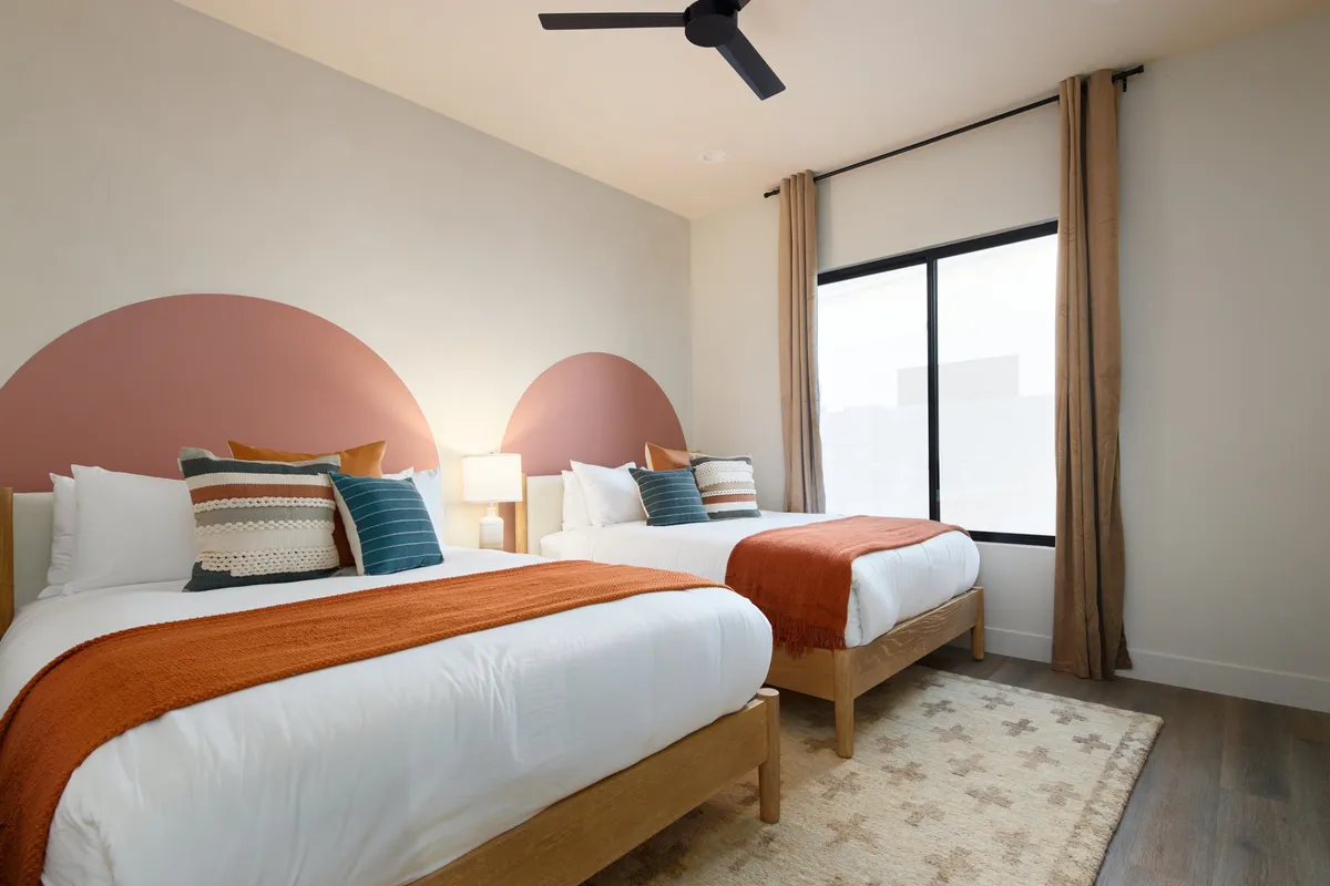 A bright bedroom featuring two beds with vibrant bedding, a large window bathing the room in natural light, and a decorative wall design. The space feels inviting and perfect for multiple guests or families traveling together.