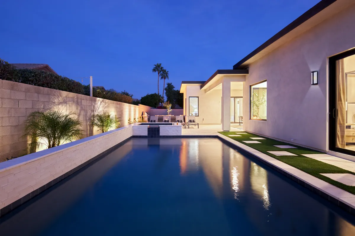 A twilight view of a luxurious backyard with a modern swimming pool, dramatic lighting, lush landscaping, and elegant patio spaces. The inviting atmosphere is perfect for evening gatherings and memorable celebrations.
